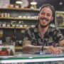 A smiling man in a cannabis shirt working behind the counter at a cannabis dispensary as a budtender
