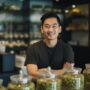 A smiling man in a black shirt behind the counter at a dispensary with jars of cannabis.