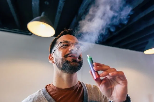 A man in a grey sweatshirt smoking a cannabis vape pen indoors