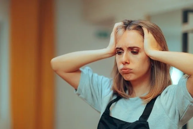 A woman in a blue shirt and an apron with her hands on her head feeling overwhelmed and stressed
