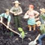 A group of five miniature farmer figurines standing around a small cannabis seedling planted in dark soil, appearing to nurture it with tools and teamwork.