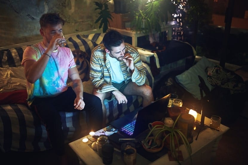 Two men relaxing on a couch in a dimly lit living room with drinks and cannabis, reflecting intentional and mindful substance use.