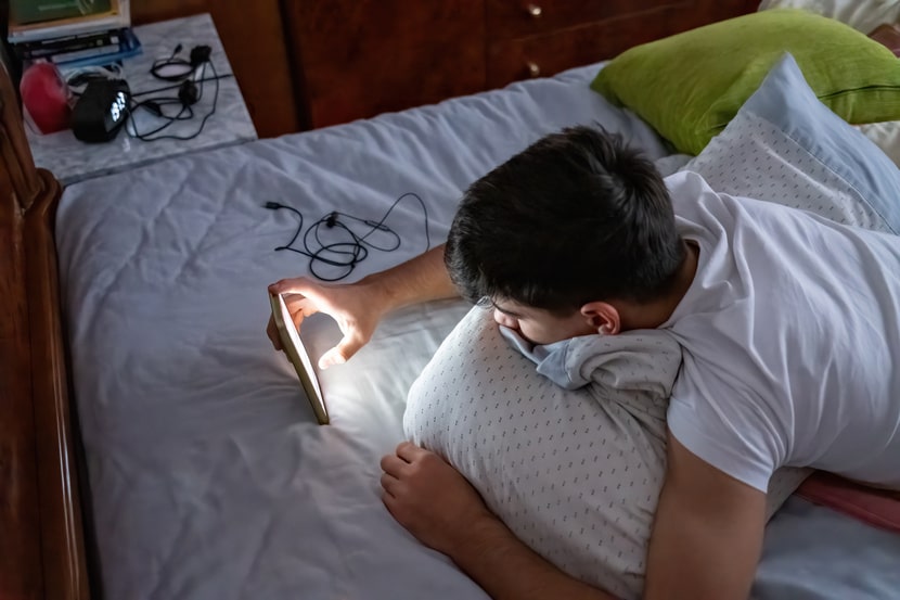 A young man lying in bed at night looking at a smartphone, illuminated by the screen in a dark bedroom.