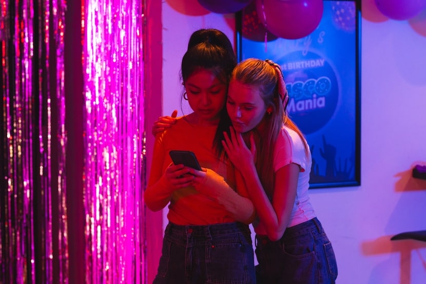 Two young women standing together at a party while looking at a phone, highlighting social media pressure and constant digital connection.