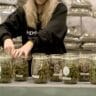 A woman in a black sweatshirt handling jars of hemp flower with large bags of hemp in the background